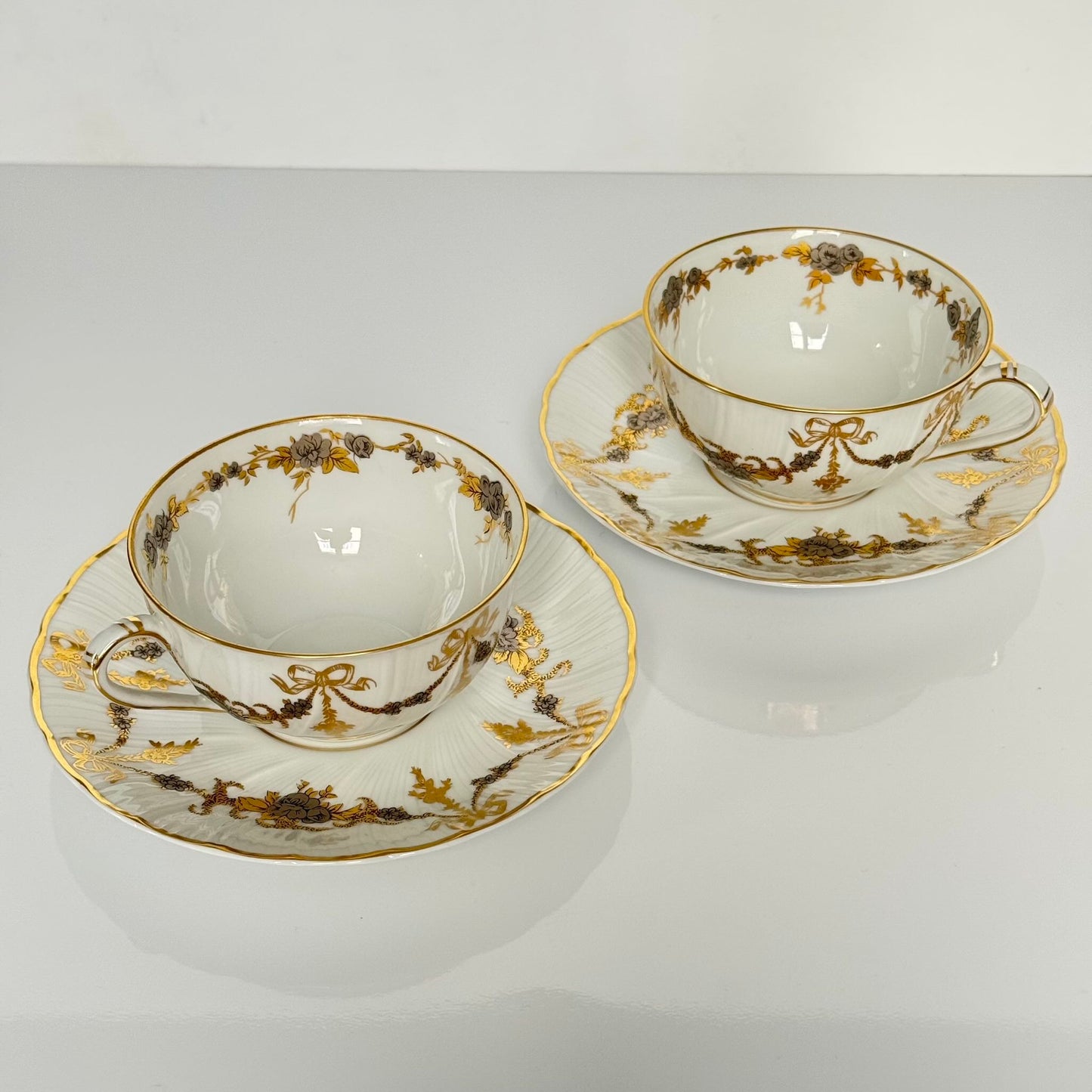 English: Pair of vintage Bernardaud Limoges tea cups and saucers with ribbon and floral garland motif, richly gilded and silvered porcelain set.; 日本語: ベルナルド リモージュ製ヴィンテージ ティーカップ&ソーサー2客セット、リボンと花のガーランド柄、金彩と銀彩装飾入りの磁器セット。; Français: Paire de tasses à thé et soucoupes vintage Bernardaud Limoges, décor de rubans/nœuds et guirlandes florales, ensemble en porcelaine richement dorée et argentée.