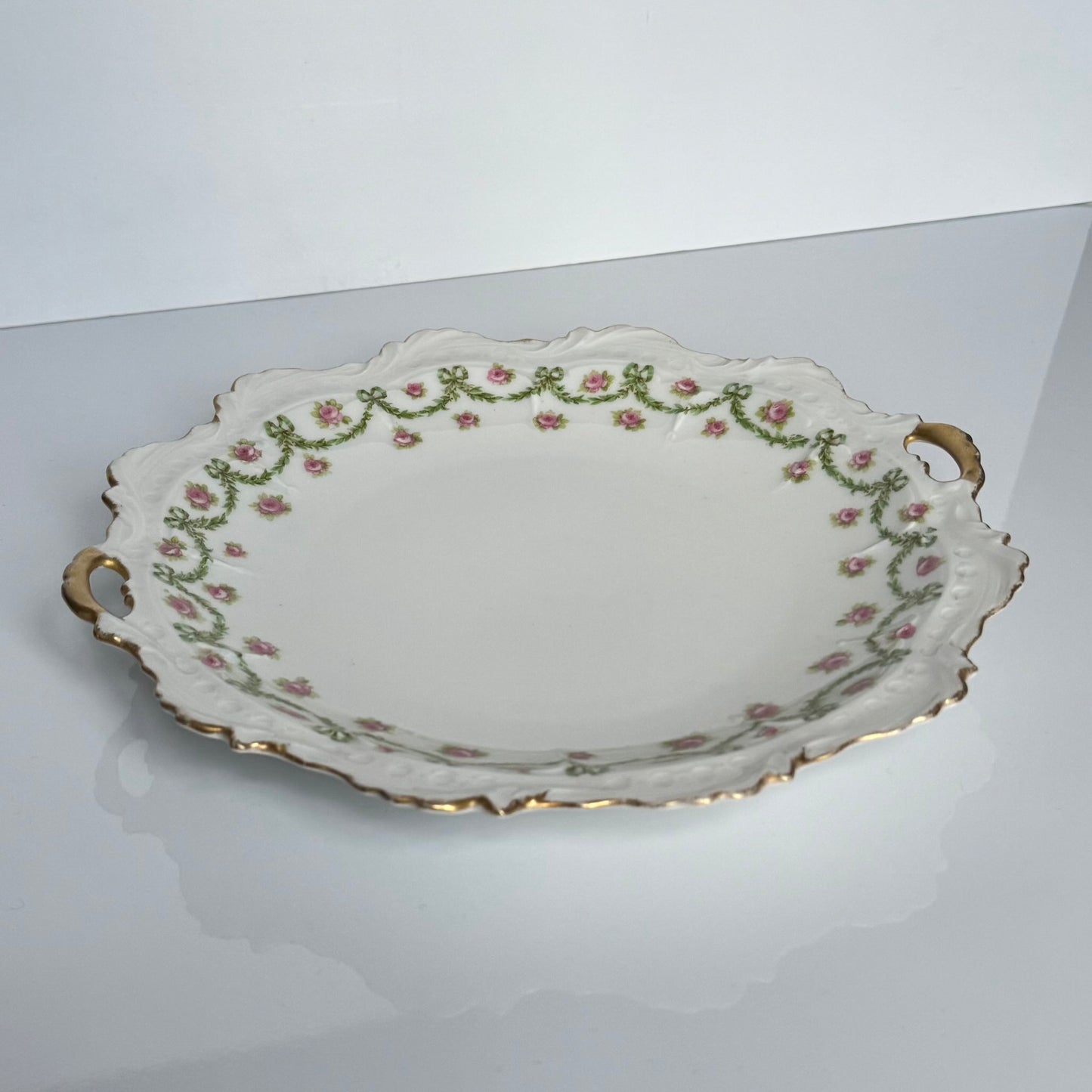 English: Limoges porcelain cake plate with scalloped edge, decorated with delicate ribbons and rose motifs in soft colors and gold accents; early 20th-century French design.; Français: Plat à gâteau en porcelaine de Limoges au bord ondulé, orné de motifs de rubans et de roses avec des touches dorées délicates ; design français du début du XXe siècle.; 日本語: リモージュ製の磁器ケーキ皿、波打つ縁と柔らかな色調のリボンとバラの装飾、繊細な金彩付き、20世紀初頭フランス製デザイン。