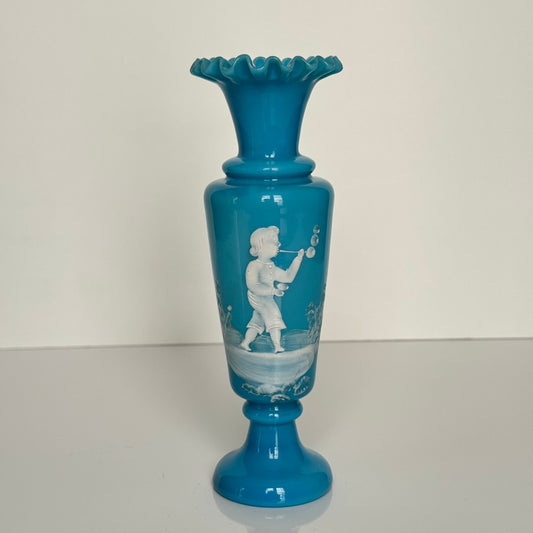 English: Blue opaline glass vase in Mary Gregory style, depicting a Victorian child blowing bubbles, hand-painted white enamel design; 日本語: メアリー・グレゴリー風の青いオパリンガラスの花瓶、シャボン玉を吹くヴィクトリア時代の子供を描いた手描き白エナメル装飾; Français: Vase en verre opalin bleu dans le style de Mary Gregory, décoré d’un enfant victorien soufflant des bulles, émaillé blanc peint à la main.