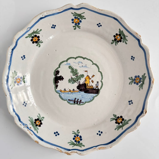 日本語: 18世紀のヌヴェール製ファイアンスプレート。風車と小さな桟橋、2隻の船が描かれた手描きのポリクローム装飾が特徴。 ; English: 18th-century Nevers faience plate, featuring a hand-painted polychrome riverside scene with a windmill, a dock, and two boats. ; Français: Assiette en faïence de Nevers du XVIIIe siècle, décorée à la main de motifs polychromes représentant une scène de rivière avec un moulin à vent, un quai et deux bateaux.