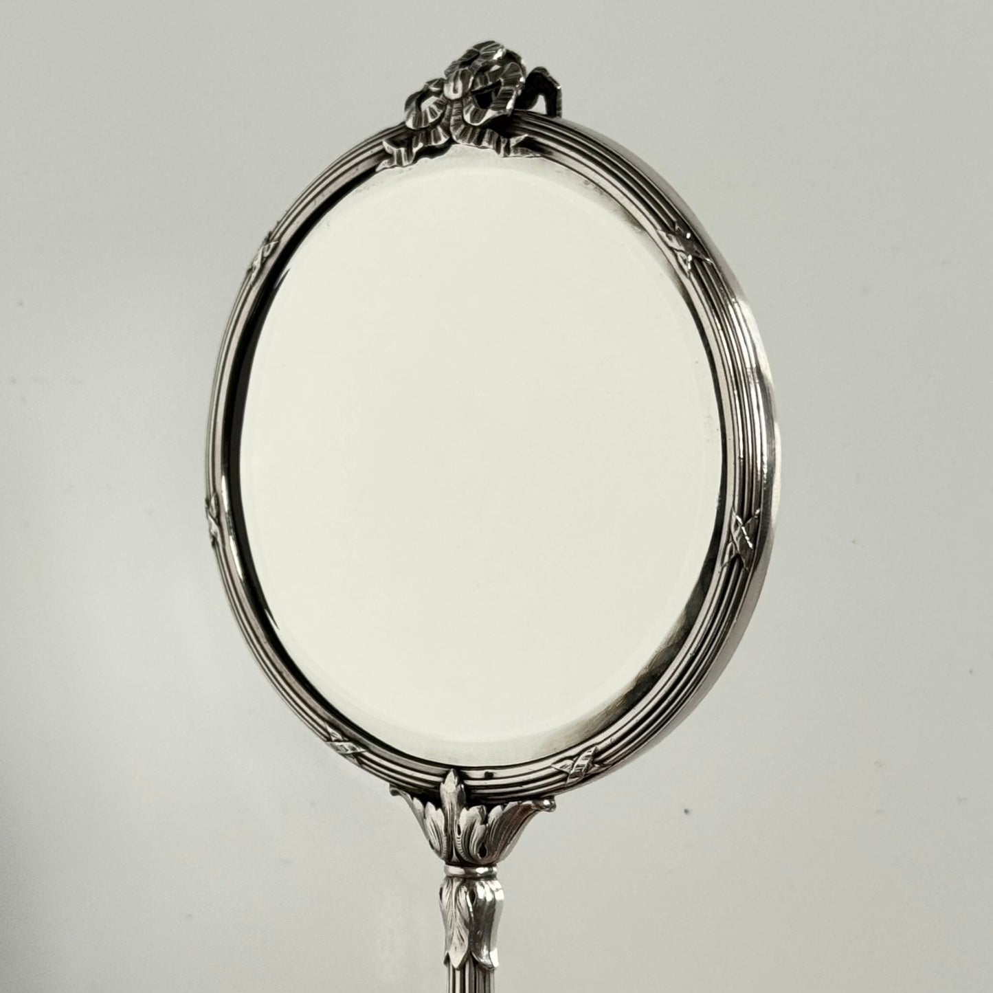 English: Silver plate hand mirror in Louis XVI style with crossed ribbons and decorative bow, late 19th to early 20th century, double-sided mirror ; 日本語: ルイ16世様式の銀メッキ手鏡、リボンと装飾的な結び目付き、19世紀末~20世紀初頭、両面鏡 ; Français: Miroir à main en métal argenté style Louis XVI avec rubans croisés et nœud, fin XIXe – début XXe siècle, miroir double face.