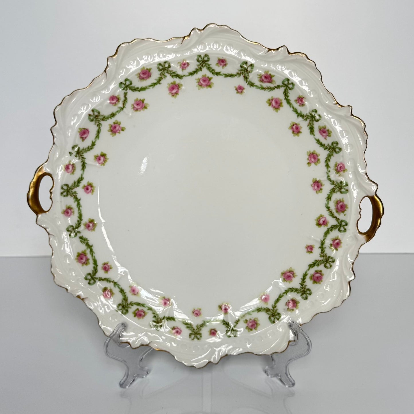 English: Limoges porcelain cake plate with scalloped edge, decorated with delicate ribbons and rose motifs in soft colors and gold accents; early 20th-century French design.; Français: Plat à gâteau en porcelaine de Limoges au bord ondulé, orné de motifs de rubans et de roses avec des touches dorées délicates ; design français du début du XXe siècle.; 日本語: リモージュ製の磁器ケーキ皿、波打つ縁と柔らかな色調のリボンとバラの装飾、繊細な金彩付き、20世紀初頭フランス製デザイン。