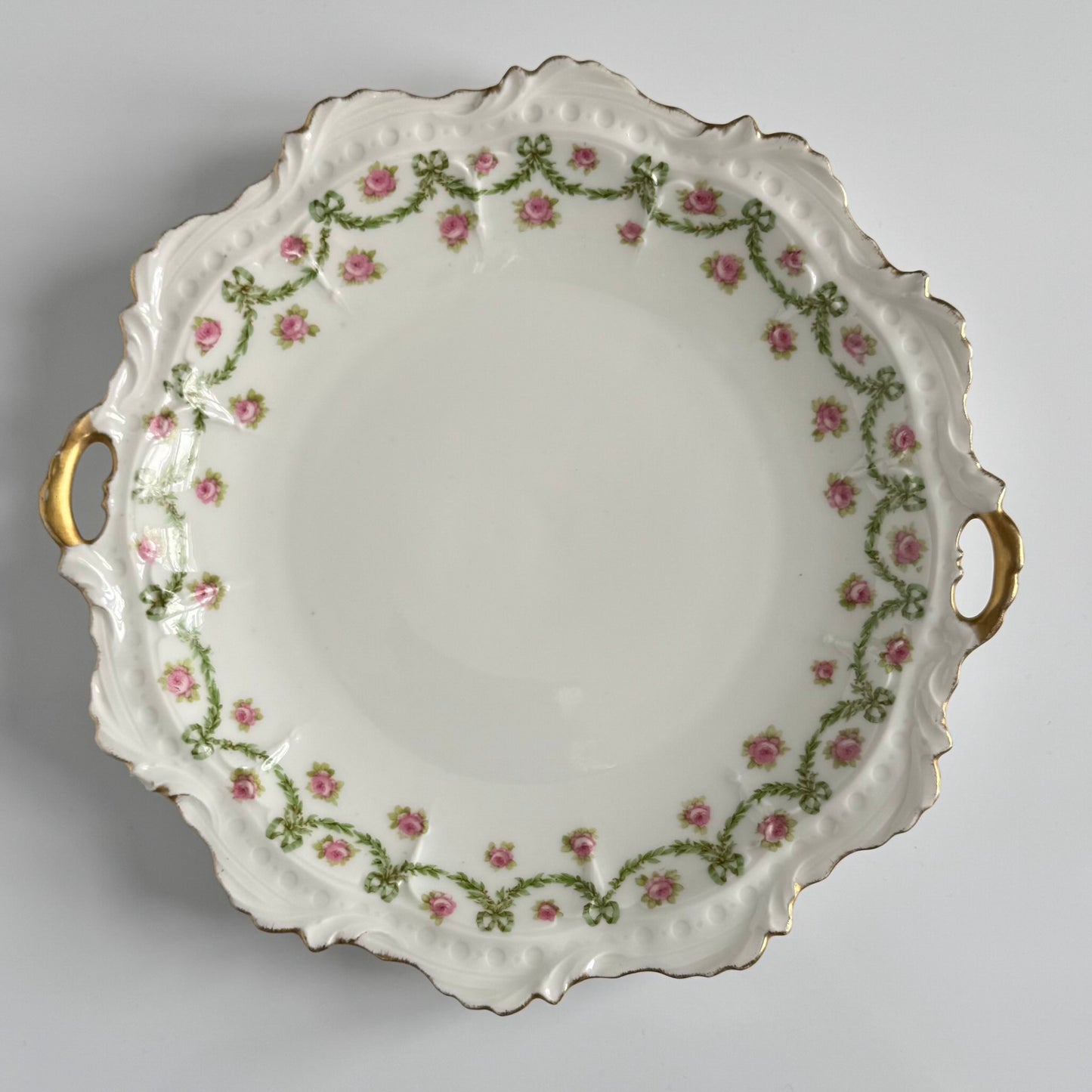 English: Limoges porcelain cake plate with scalloped edge, decorated with delicate ribbons and rose motifs in soft colors and gold accents; early 20th-century French design.; Français: Plat à gâteau en porcelaine de Limoges au bord ondulé, orné de motifs de rubans et de roses avec des touches dorées délicates ; design français du début du XXe siècle.; 日本語: リモージュ製の磁器ケーキ皿、波打つ縁と柔らかな色調のリボンとバラの装飾、繊細な金彩付き、20世紀初頭フランス製デザイン。