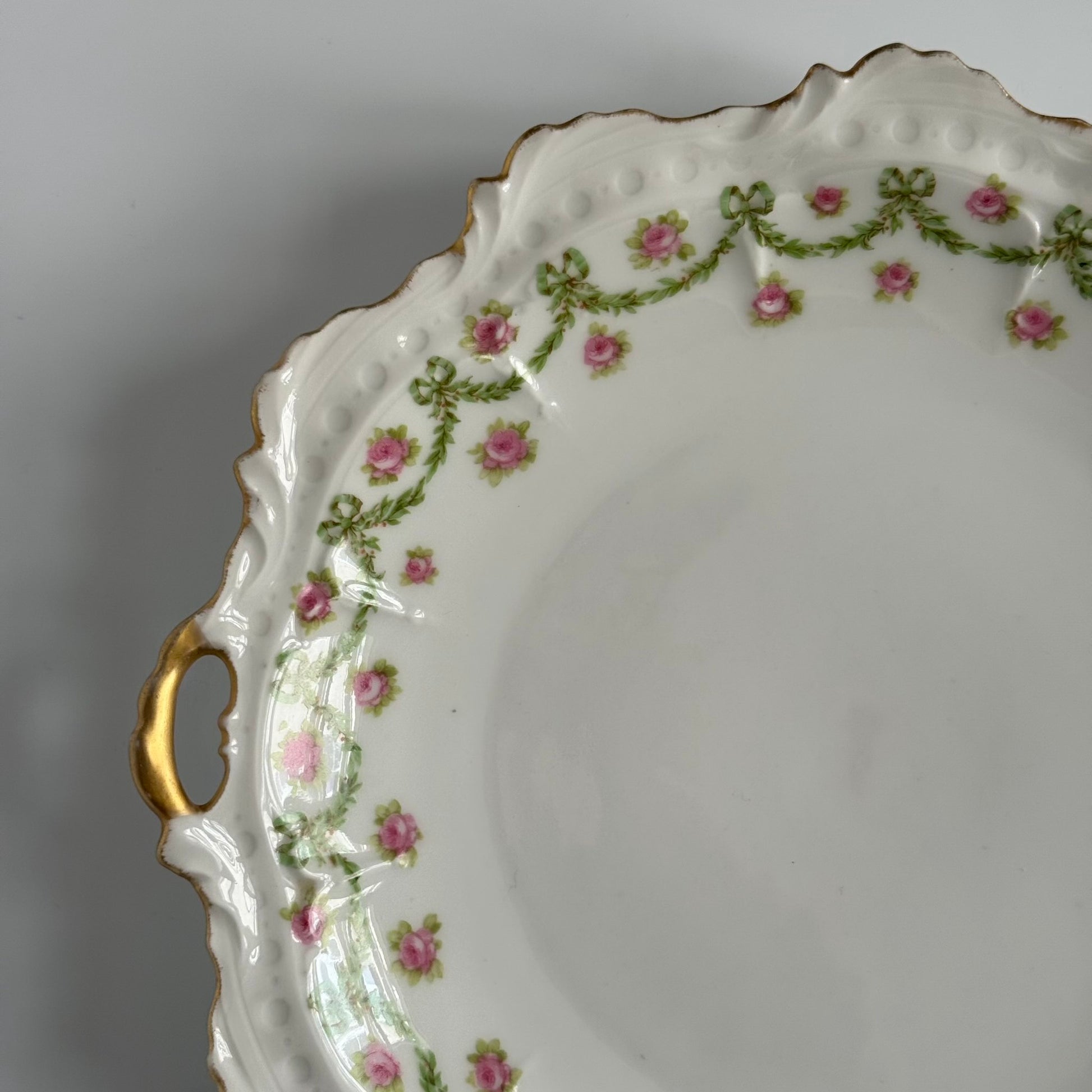 English: Limoges porcelain cake plate with scalloped edge, decorated with delicate ribbons and rose motifs in soft colors and gold accents; early 20th-century French design.; Français: Plat à gâteau en porcelaine de Limoges au bord ondulé, orné de motifs de rubans et de roses avec des touches dorées délicates ; design français du début du XXe siècle.; 日本語: リモージュ製の磁器ケーキ皿、波打つ縁と柔らかな色調のリボンとバラの装飾、繊細な金彩付き、20世紀初頭フランス製デザイン。