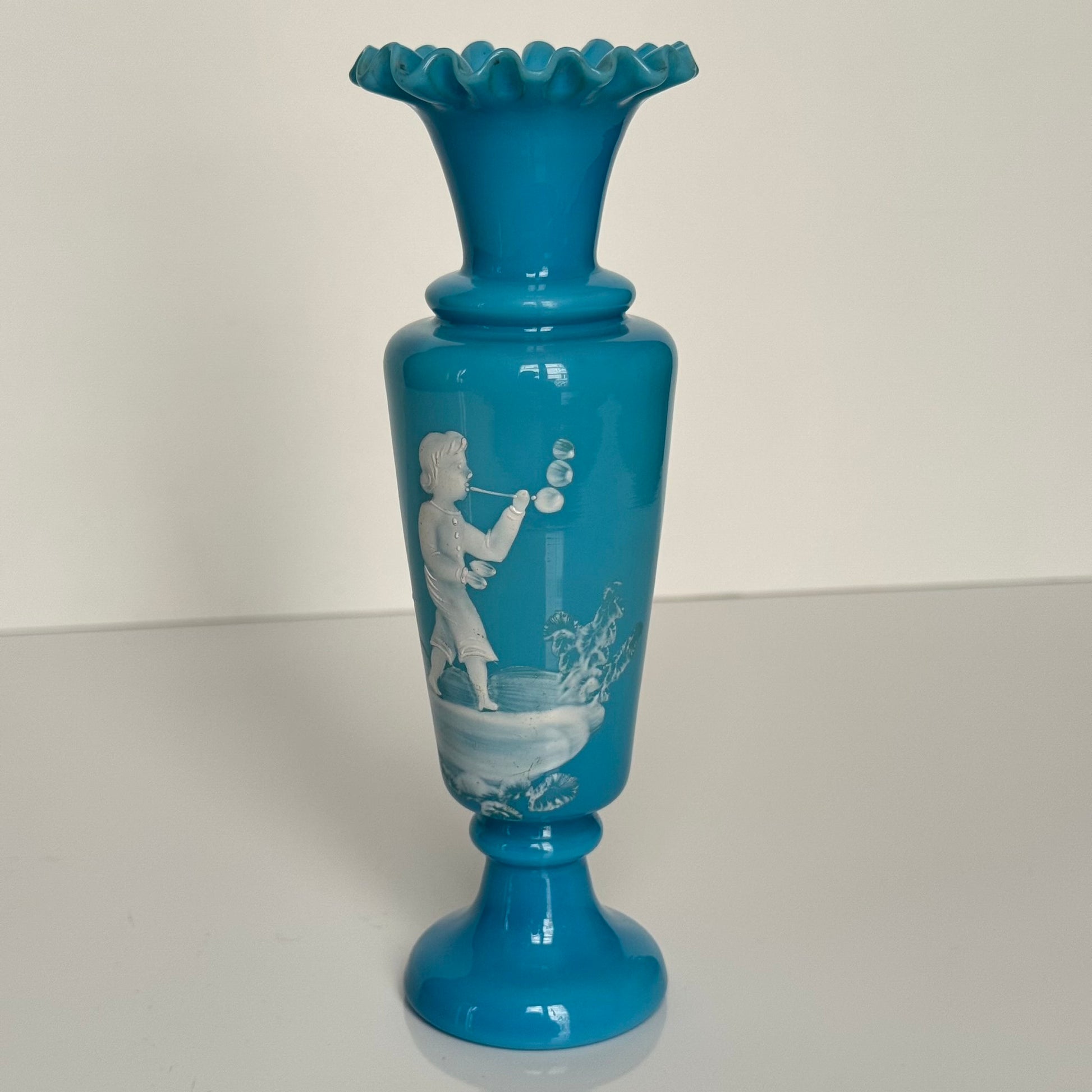 English: Blue opaline glass vase in Mary Gregory style, depicting a Victorian child blowing bubbles, hand-painted white enamel design; 日本語: メアリー・グレゴリー風の青いオパリンガラスの花瓶、シャボン玉を吹くヴィクトリア時代の子供を描いた手描き白エナメル装飾; Français: Vase en verre opalin bleu dans le style de Mary Gregory, décoré d’un enfant victorien soufflant des bulles, émaillé blanc peint à la main.