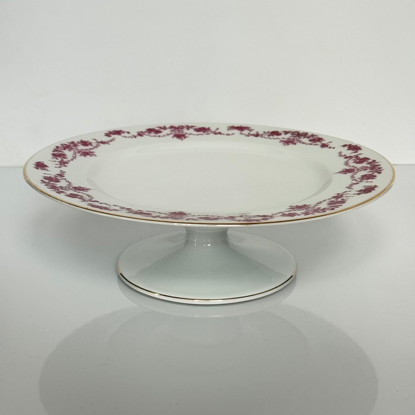 English: Antique Pirkenhammer porcelain cake stand decorated with ribbon and floral garland motif in pink and violet, early 20th century. ; Français : Présentoir à gâteau en porcelaine Pirkenhammer, décor de rubans et guirlandes florales rose/violet, début XXᵉ siècle. ; 日本語： ピルケンハンマー製アンティークケーキスタンド、ピンクパープルのリボンと花のガーランド装飾、20世紀初頭。