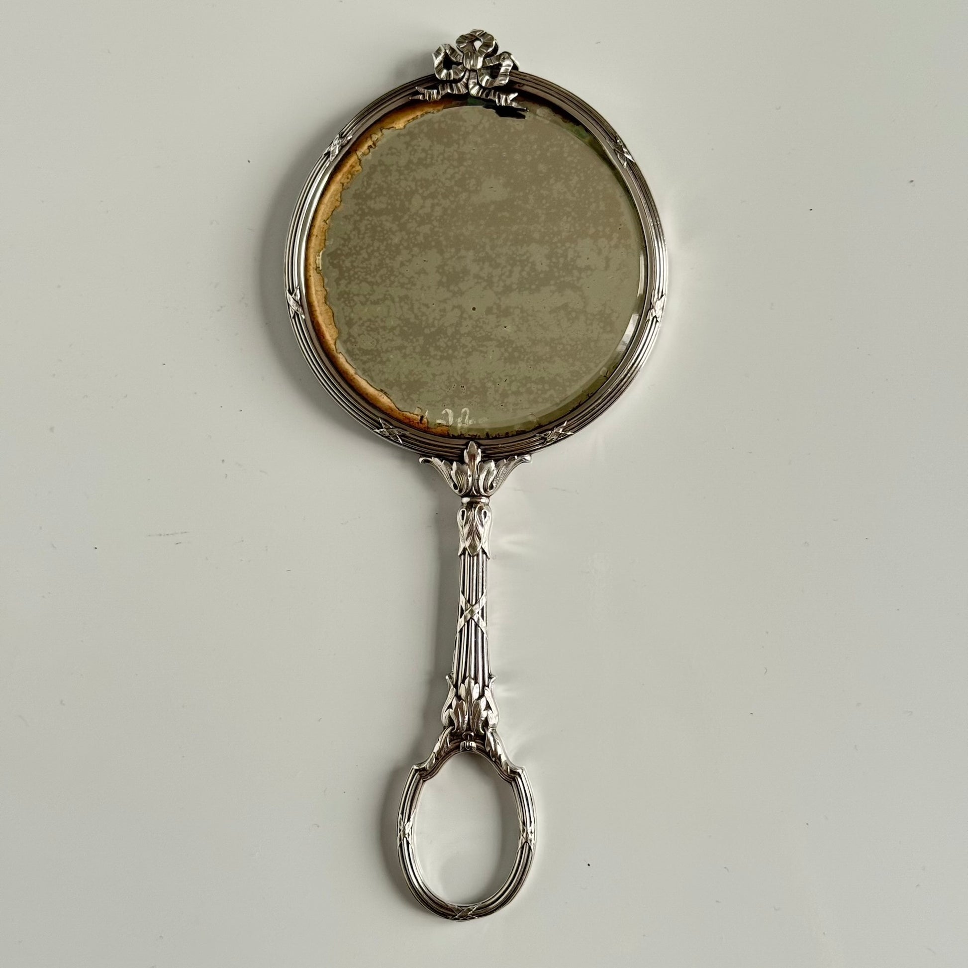 English: Silver plate hand mirror in Louis XVI style with crossed ribbons and decorative bow, late 19th to early 20th century, double-sided mirror ; 日本語: ルイ16世様式の銀メッキ手鏡、リボンと装飾的な結び目付き、19世紀末～20世紀初頭、両面鏡 ; Français: Miroir à main en métal argenté style Louis XVI avec rubans croisés et nœud, fin XIXe – début XXe siècle, miroir double face.