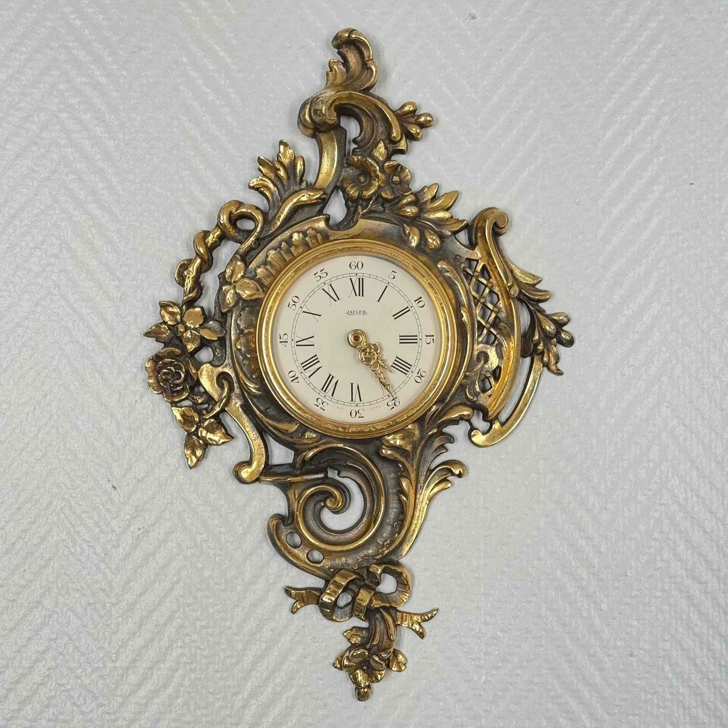English: Vintage French wall clock by Jaeger in gilded bronze, Rococo style with scrolls, flowers, ribbons, and serpent motif, 1950s–60s. ; 日本語: ジャガー製のフランス製ヴィンテージ壁掛け時計、1950〜60年代、ギルドブロンズ製、ロココ様式のスクロールや花、リボン、蛇の装飾付き。 ; Français : Pendule murale vintage française par Jaeger en bronze doré, style rococo avec volutes, fleurs, rubans et motif de serpent, années 1950–60.